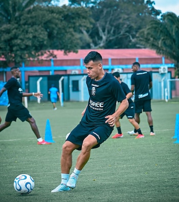 Jugador de fútbol de Blooming con uniforme negro y botines claros entrenando en césped artificial, junto a un balón. Otros jugadores y conos azules en el fondo