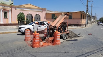 Dos operarios con cascos naranjas manipulan maquinaria de limpieza junto a un buzón de alcantarillado cercado con conos y mallas de seguridad