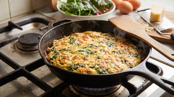 Una sartén de hierro sobre una cocina contiene una tortilla dorada, junto a un bol con hojas verdes, huevos, queso y un rallador.