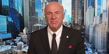 Hombre calvo de mediana edad con traje oscuro y corbata blanca, expresión seria, mirando a cámara. Fondo virtual de rascacielos urbanos y edificios bajos