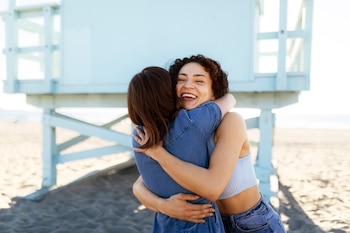 Dos chicas se dan un abrazo en la playa, con una caseta de color azul tras de ellas sobre la arena