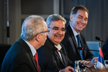 Tres hombres en traje sentados en una mesa, dos de ellos sonríen, con micrófonos y una pequeña bandera de franjas roja, azul y blanca