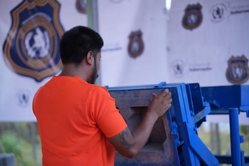 Hombre de espaldas con camiseta naranja, cabello oscuro y barba. Manos sobre una estructura metálica azul. Fondo con banderas blancas y escudos borrosos