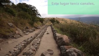 Un camino de tierra con muros de piedra a los lados, rodeado de rocas y vegetación en colinas; logo de Cultura y texto en un recuadro verde que dice 'Este lugar tenía canales,'