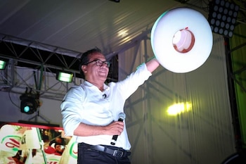 Roberto Sánchez sonríe en un escenario, sosteniendo un micrófono y un objeto circular blanco, con luces de escenario y estructura metálica de fondo