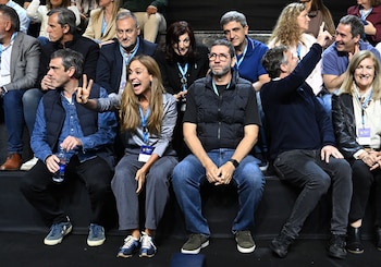 Grupo de personas sentadas en una fila. Un hombre hace la señal de victoria, una mujer ríe a carcajadas. El hombre CREW M lleva gafas y chaleco oscuro