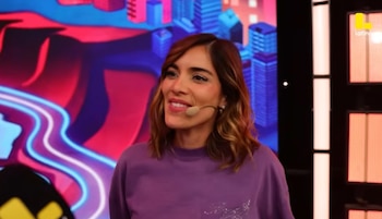 Korina Rivadeneira, con cabello castaño claro ondulado y una sudadera morada, sonríe ligeramente con un micrófono en la mejilla, frente a un fondo colorido de ciudad