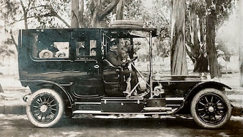 Fotografía en blanco y negro de un automóvil antiguo con un chofer y pasajeros en su interior, estacionado junto a varios árboles