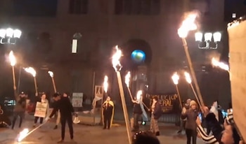 Personas sostienen antorchas con fuego en una plaza nocturna frente a pancartas con texto y faroles de calle encendidos