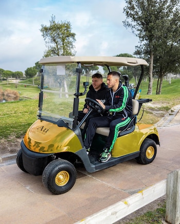 Dos hombres en un golf cart dorado con logo "Au Vodka" en un campo de golf. Hay árboles, césped, un lago y un camino pavimentado