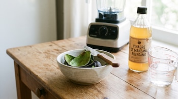 Un bol de cerámica con cáscaras de aguacate y un cuchillo pequeño en una mesa de madera. Una botella de vinagre de manzana y un vaso medidor con agua. Al fondo, una licuadora.