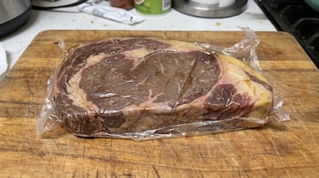 Un corte de carne roja con grasa blanca y amarillenta, envasado en plástico transparente, sobre una tabla de cortar de madera.