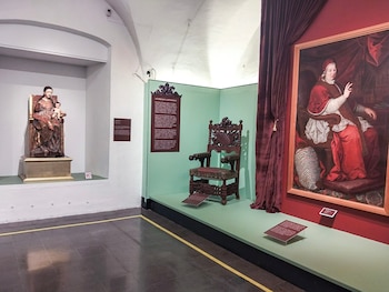 Una escultura de la Virgen con el Niño, un sillón de madera y un cuadro al óleo de un religioso en un museo
