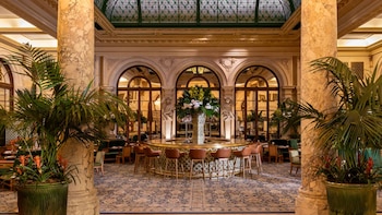 Un salón con columnas de mármol, una barra circular con taburetes, plantas de palmera en macetas grandes y arcos acristalados al fondo