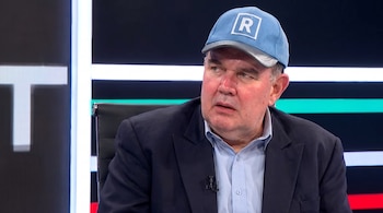 Un hombre de mediana edad con gorra azul claro con una R, camisa celeste y saco azul oscuro, sentado en una silla negra frente a un fondo oscuro con luces.