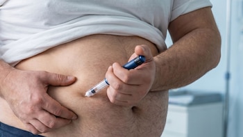 Un hombre se inyecta con una pluma de inyección en el abdomen. Viste una camiseta blanca, y sus manos sujetan la piel del abdomen y la pluma de inyección