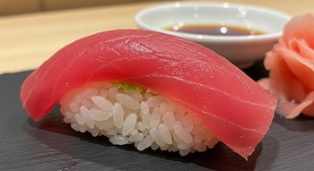 Maguro nigiri con atún rojo sobre arroz blanco en plato oscuro. Al fondo, salsa de soya y jengibre encurtido.