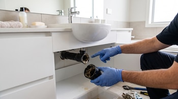 Una persona con guantes azules sostiene un tramo de tubería de plástico con residuos acumulados en el interior bajo un lavabo.