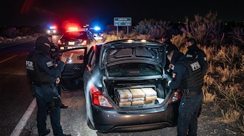 Agentes encapuchados con chalecos tácticos revisan el maletero de un vehículo iluminado por patrullas en una carretera de noche.