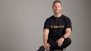 Hombre caucásico sonriente con barba, camiseta negra con el logo dorado de Binance, pantalón negro y reloj oscuro, sentado en un taburete