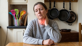 Mujer sentada en una cocina con la tiroides resaltada en rojo en su cuello. Detrás hay utensilios de plástico coloridos y sartenes antiadherentes colgadas.