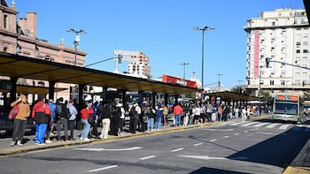 Largas filas de personas esperan en paradas de colectivo amarillas en una calle urbana. Un autobús verde se acerca. Edificios y cielo azul despejado al fondo