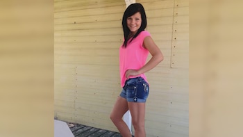 Una mujer joven con cabello oscuro y una camiseta rosa fuerte sonríe mientras posa de pie frente a una pared de tablones de madera clara