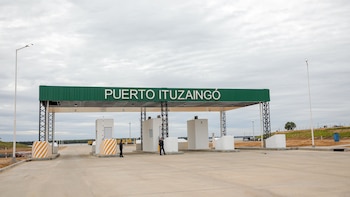 Cartel verde con "PUERTO ITUZAINGÓ", estructura metálica, garitas blancas, dos hombres, barreras, postes de luz y una carretera de concreto
