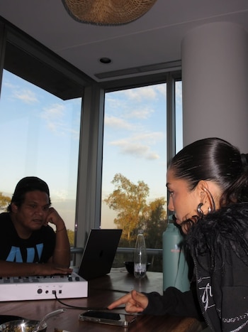 Una mujer con chaqueta negra y cabello recogido señala un teléfono. A su lado, un hombre con gorra frente a un controlador MIDI y un portátil. Mesa de madera con vista a árboles y cielo