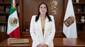 Ana Miriam Ramos Villareal, Alcaldesa Sustituta de Culiacán, de pie en una oficina con blazer blanco. Bandera de México y escudo de Culiacán al fondo.