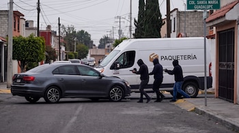 Tres individuos encapuchados con pistolas se aproximan a una camioneta blanca de paquetería, mientras un coche sedán gris detiene su marcha en una calle urbana.