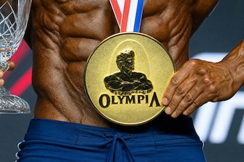 Torso muscular de un hombre que viste pantalón azul y sostiene una medalla dorada de Olympia y un trofeo de cristal.
