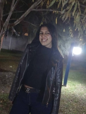 Una mujer con chaqueta negra de abrigo y cabello oscuro sonríe de noche cerca de un árbol y un farol encendido