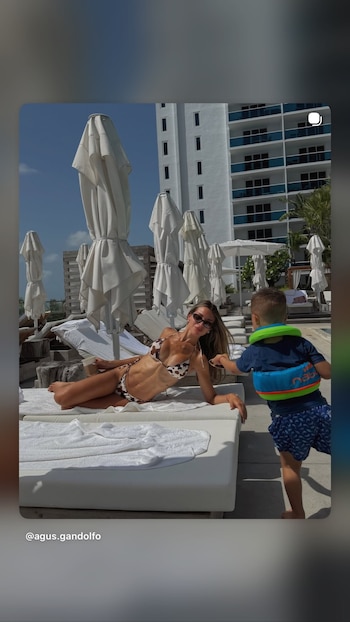 Mujer en bikini y lentes de sol recostada en reposera blanca al aire libre, con niño en chaleco de flotación azul y verde