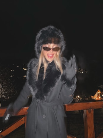 Una mujer sonriente con abrigo negro, gorro de piel, gafas de sol y guantes negros junto a una baranda de madera de noche