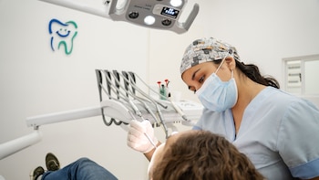 Una dentista con mascarilla, guantes y gorro sanitario examina la boca de un paciente. Equipo dental, lámpara, mangueras y un logo de muela en la pared