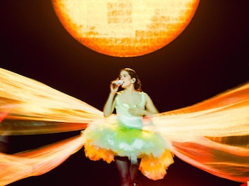 Cantante Rosalía con micrófono, top blanco, falda de tul verde y naranja. Fondo oscuro, gran círculo luminoso naranja y tiras de tela naranjas extendidas
