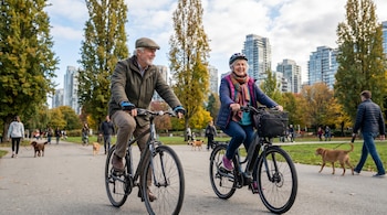 Un hombre y una mujer mayores en bicicleta en un parque. El hombre con gorra y chaqueta, la mujer con casco y bufanda. Árboles altos y edificios urbanos detrás.