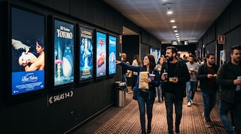 Una mujer y un hombre caminan por el pasillo de un cine con palomitas de maíz y bebidas frente a carteles de películas.