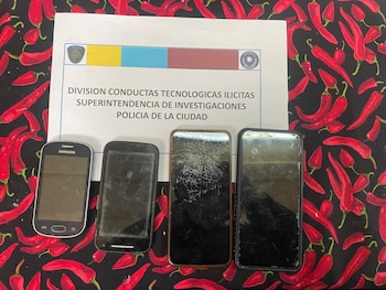 Cuatro teléfonos móviles, uno con pantalla rota, sobre una tela oscura con estampado de pimientos rojos, y un documento oficial de la Policía de la Ciudad