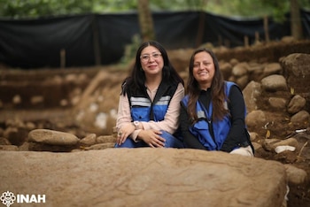 Dos mujeres sonríen frente a la cámara en un sitio arqueológico con tierra y piedras. Hay una losa de piedra labrada en primer plano. Logotipo INAH