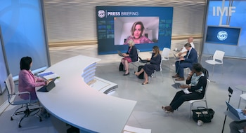 Sala de conferencias, pantalla grande con mujer en videollamada, personas sentadas con laptops, mesa curva, logo del FMI, sillas