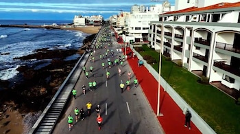 Vista aérea de una avenida costera con muchas personas en ropa deportiva. La avenida incluye asfalto y una senda roja. Al lado, edificios, playa, rocas y mar