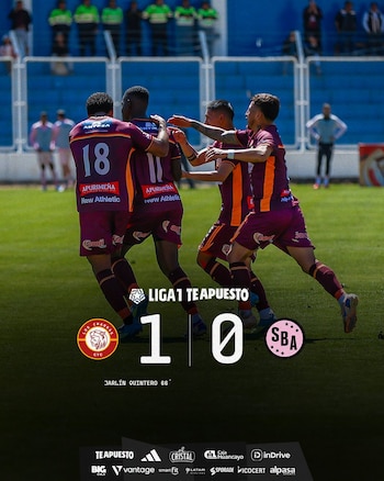 Jugadores de fútbol con camisetas granates y naranjas celebran en un campo de césped. En la parte inferior, un marcador digital muestra el resultado 1-0