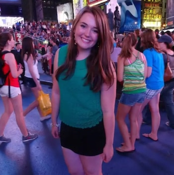 Una mujer de cabello castaño y blusa verde sonríe de pie en Times Square entre transeúntes y letreros luminosos