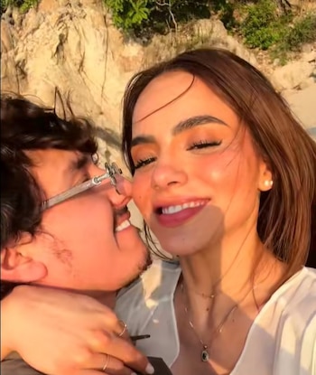 Primer plano de un hombre con gafas y una mujer sonriente abrazados. De fondo, una playa con rocas y vegetación bajo luz dorada