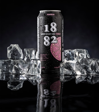 Lata negra de bebida 1882 Fermelo con sabor a fernet y pomelo, cubierta de condensación, rodeada de cubos de hielo sobre una superficie reflectante y fondo oscuro