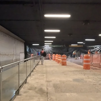 Pasillo en remodelación de la Línea 2 del Metro CDMX con barandilla metálica, suelo de concreto, barriles naranjas de construcción y operarios al fondo