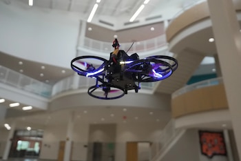 Dron negro volando en el interior de un edificio moderno con barandales, escaleras y luces de techo. El dron tiene hélices y luces azules