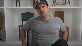 Un hombre con gorra y camiseta gris se sienta en una silla de mimbre, mirando al frente. Detrás, una estantería blanca con objetos decorativos